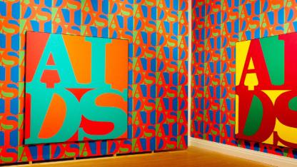 Blick in die Ausstellung: An zwei rot-blau-grün gemusterten Wänden hängt jeweils ein Gemälde mit dem AIDS-Logo in GRoßbuchstaben, einmal in Grün auf Rot, einmal in Rot auf gelb-grünem Hintergrund