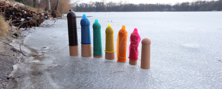 Es ist ein zugefrorener See zu sehen. Auf dem See stehen sieben Holzmodelle von Penissen, die von links nach rechts kleiner werden. Auf den sechs linken Holzmodellen sind Kondome drauf. Von links nach rechts in lila, blau, grün, gelb, orange und pink. Das Holzmodell ganz rechts hat kein Kondom drauf. 