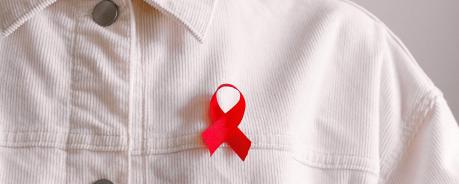 Das Bild zeigt einen Menschen mit einer weißen Cordjacke und einem Anstecker mit der roten Aidsschleife. 