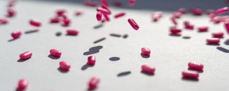 Bild zeigt einen weißen Tisch, auf denen viele längliche, pinke Pillen liegen. Manche Pillen sind noch in der Luft, als hätte sie jemand auf den Tisch geworfen.