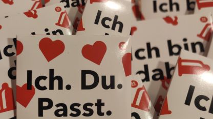 Viele weiße Kondomumschläge mit roten Herzen und Kondomen aufgedruckt. In schwarzer Schrift steht auf den Umschlägen "Ich bin dabei." oder "Ich. Du. Passt."